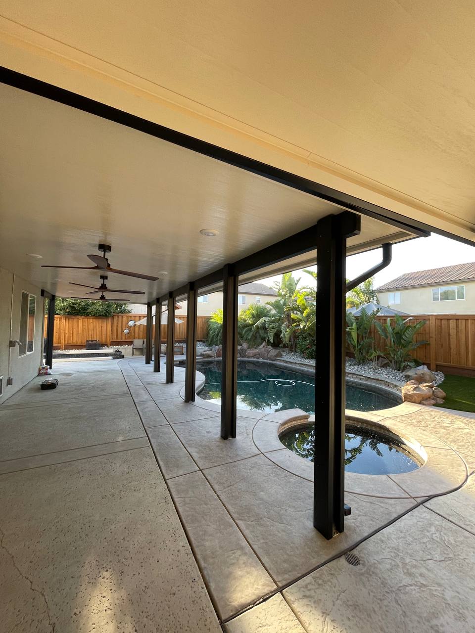 Full-length custom attached solid insulated aluminum patio cover with dark bronze posts, integrated modern ceiling fans, and downspouts shading a poolside deck and spa in Elk Grove, CA.